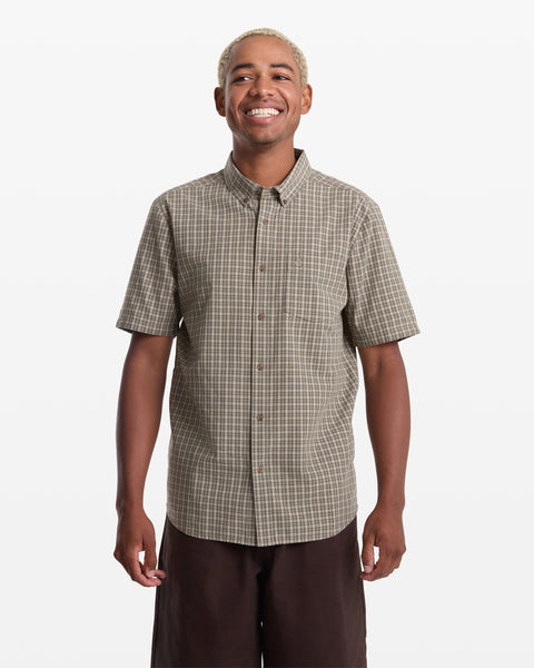 A person with short blond hair smiles in a Volcom Men's Brewster Short Sleeve brown plaid shirt and dark pants, standing against a plain white background.