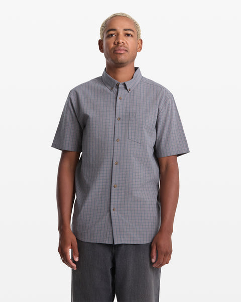 A person with short blond hair wears the Volcom Men's Brewster Short Sleeve shirt—a gray, checkered, button-up with a chest pocket—standing against a plain white background.
