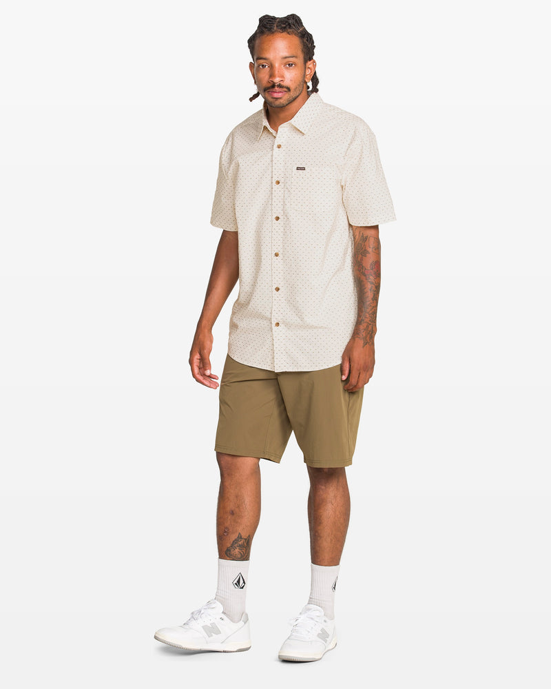 A man with braided hair wears the Volcom Men's Bankstone II Short Sleeve Tee, olive green shorts, white socks, and white sneakers as he stands against a plain white background.