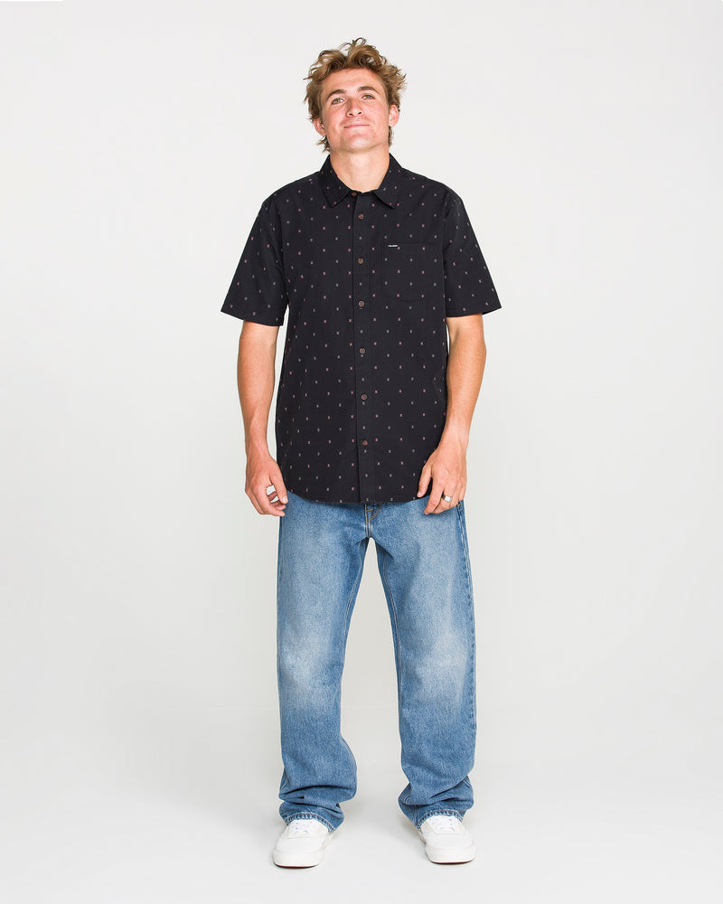 A person stands against a plain white background, wearing the Volcom Men's Bankstone Short Sleeve Shirt, featuring a small pattern, paired with light blue jeans and white sneakers. The classic fit shirt is crafted from soft cotton-poly fabric for all-day comfort and style.