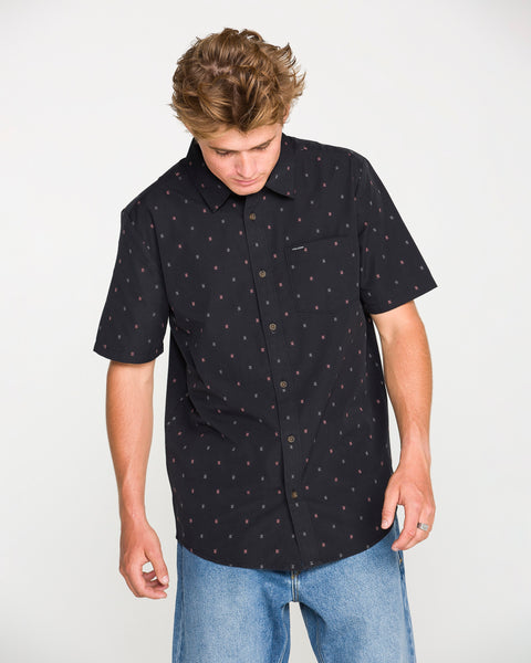 A person wearing the Volcom Men's Bankstone Short Sleeve Shirt paired with light blue jeans looks down with both hands at their sides against a plain background.