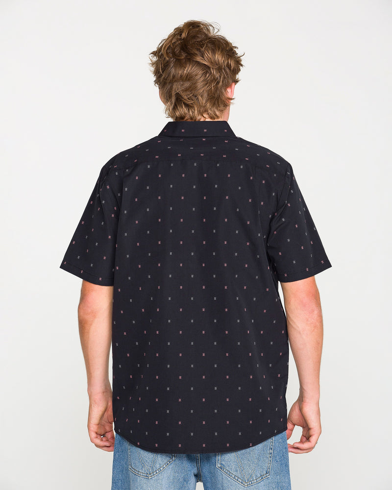 A person with light brown hair is shown from the back against a plain white background, wearing the Volcom Men's Bankstone Short Sleeve Shirt—a black button-up featuring a small red dotted pattern—paired with blue jeans.