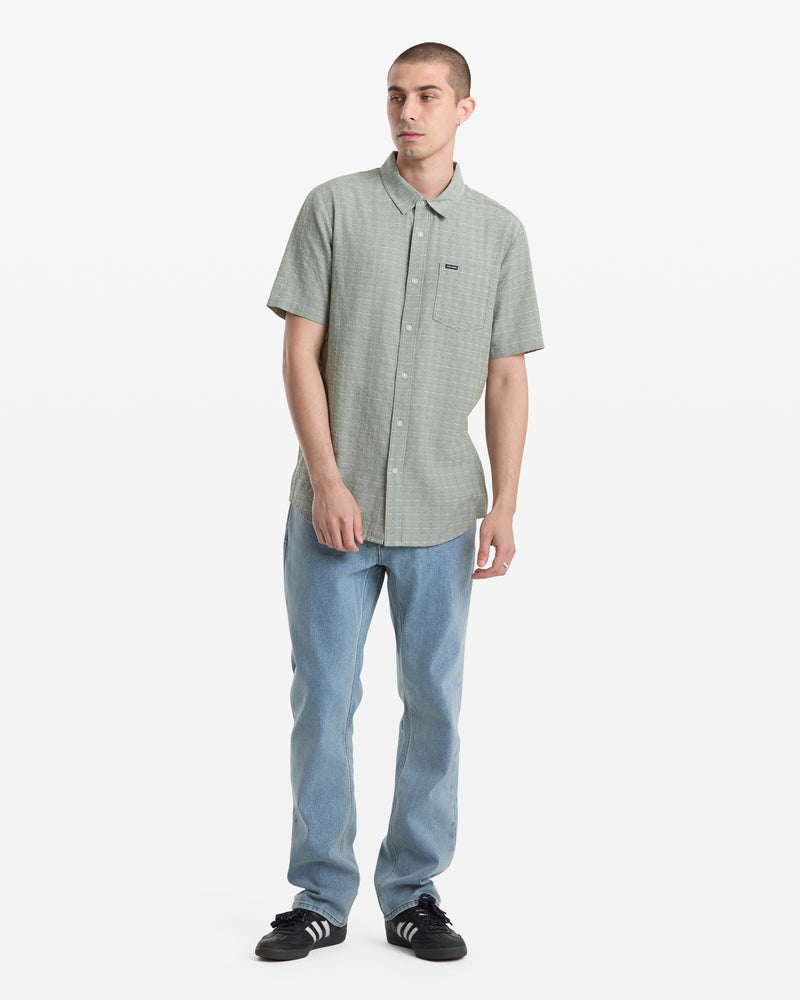 A man stands against a plain white background, wearing the Volcom Men's Bankstone II Short Sleeve shirt in light green, paired with light blue jeans and black sneakers with white stripes.
