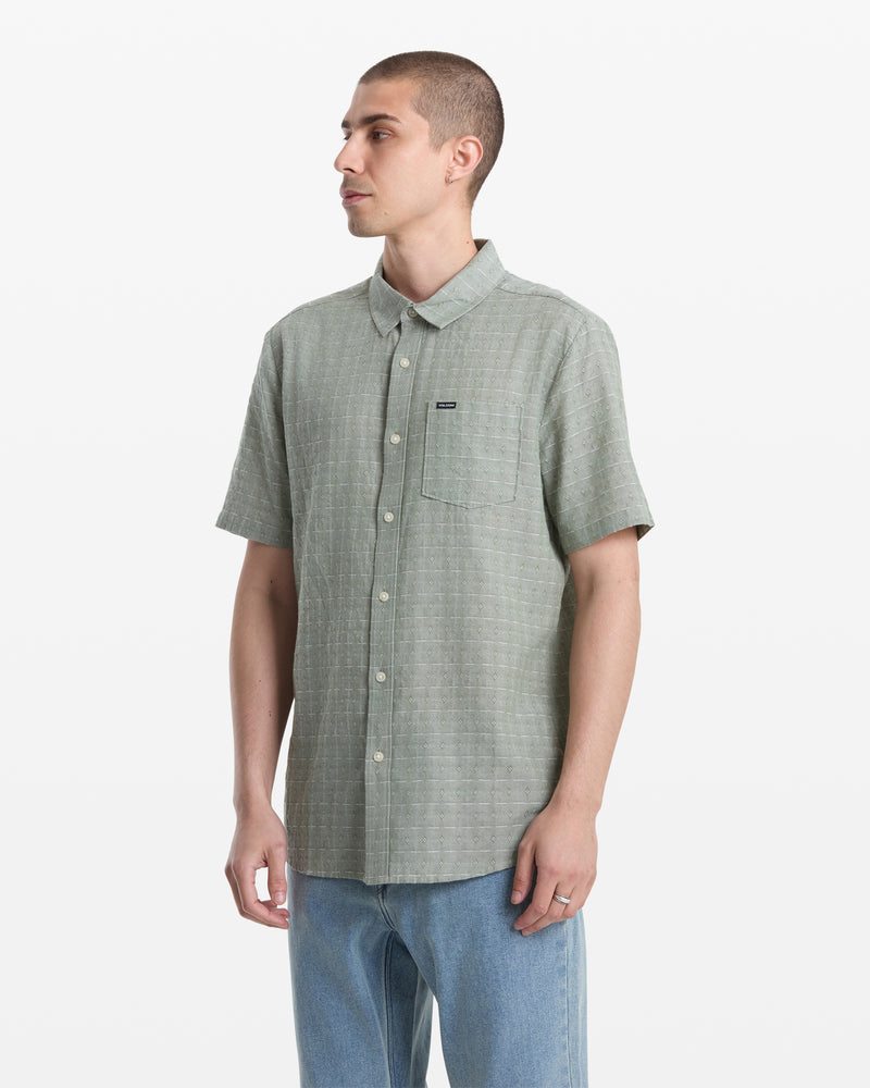 A man with short hair wears the Volcom Men's Bankstone II Short Sleeve shirt in light green with a chest pocket and yarn-dyed dobby texture, paired with light blue jeans, standing against a plain white background.