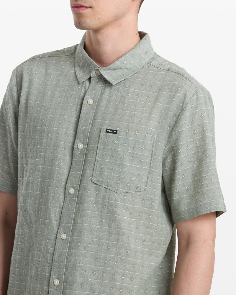 A man wears the Volcom Men's Bankstone II Short Sleeve shirt in light green with a classic fit, yarn-dyed dobby texture, chest pocket, and a small black rectangular tag. The image shows him from shoulders to mid-torso.