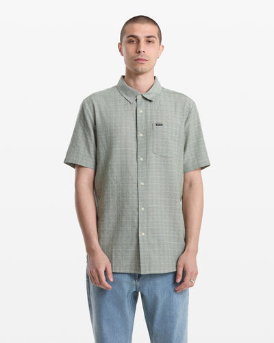 A man with short hair wears the Volcom Men's Bankstone II Short Sleeve shirt in light green, featuring a yarn-dyed dobby texture, chest pocket, and blue jeans as he stands facing forward against a plain white background.