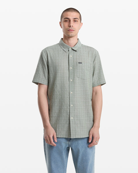A man with short hair wears the Volcom Men's Bankstone II Short Sleeve shirt in light green, featuring a yarn-dyed dobby texture, chest pocket, and blue jeans as he stands facing forward against a plain white background.