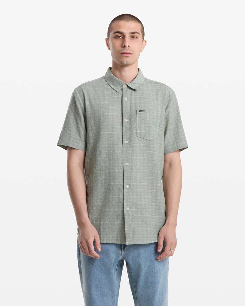 A man with short hair wears the Volcom Men's Bankstone II Short Sleeve shirt in light green, featuring a yarn-dyed dobby texture, chest pocket, and blue jeans as he stands facing forward against a plain white background.