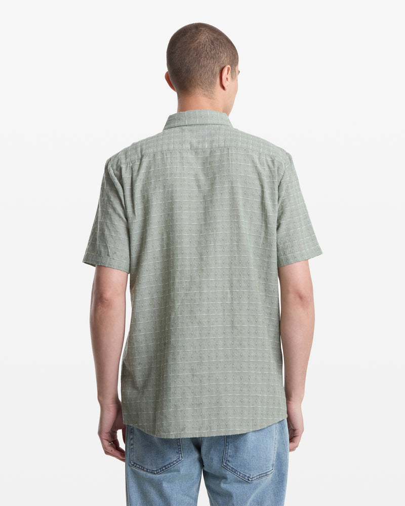 Seen from behind on a plain white background, a person with short hair wears the Volcom Men's Bankstone II Short Sleeve shirt in light green, featuring a classic fit and subtle dobby texture, paired with light blue jeans.
