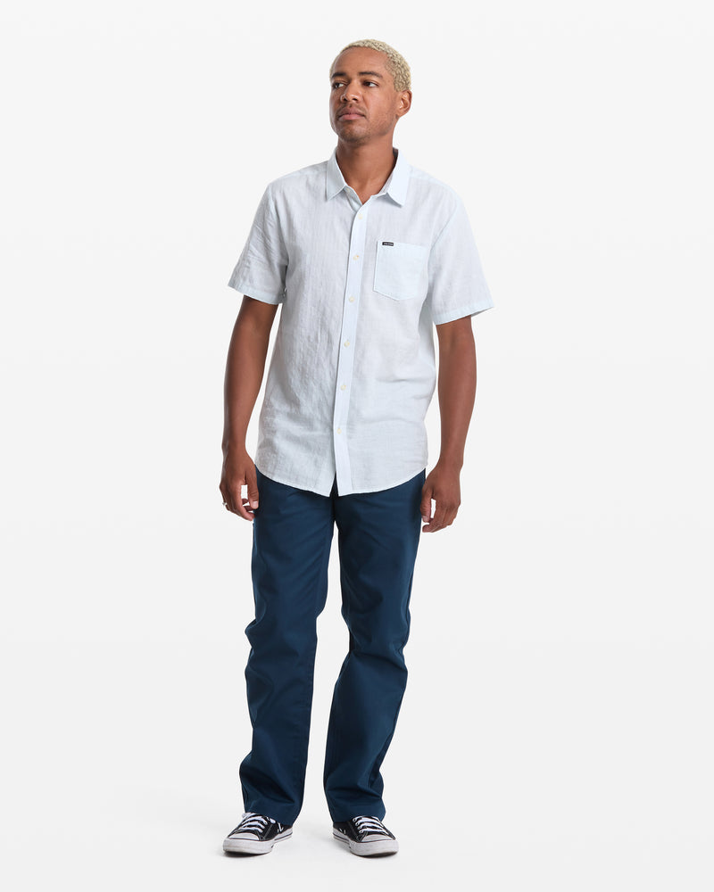 A man with short blond hair wears the Volcom Men's Bankstone II Short Sleeve shirt, blue pants, and dark sneakers, standing against a plain white background.