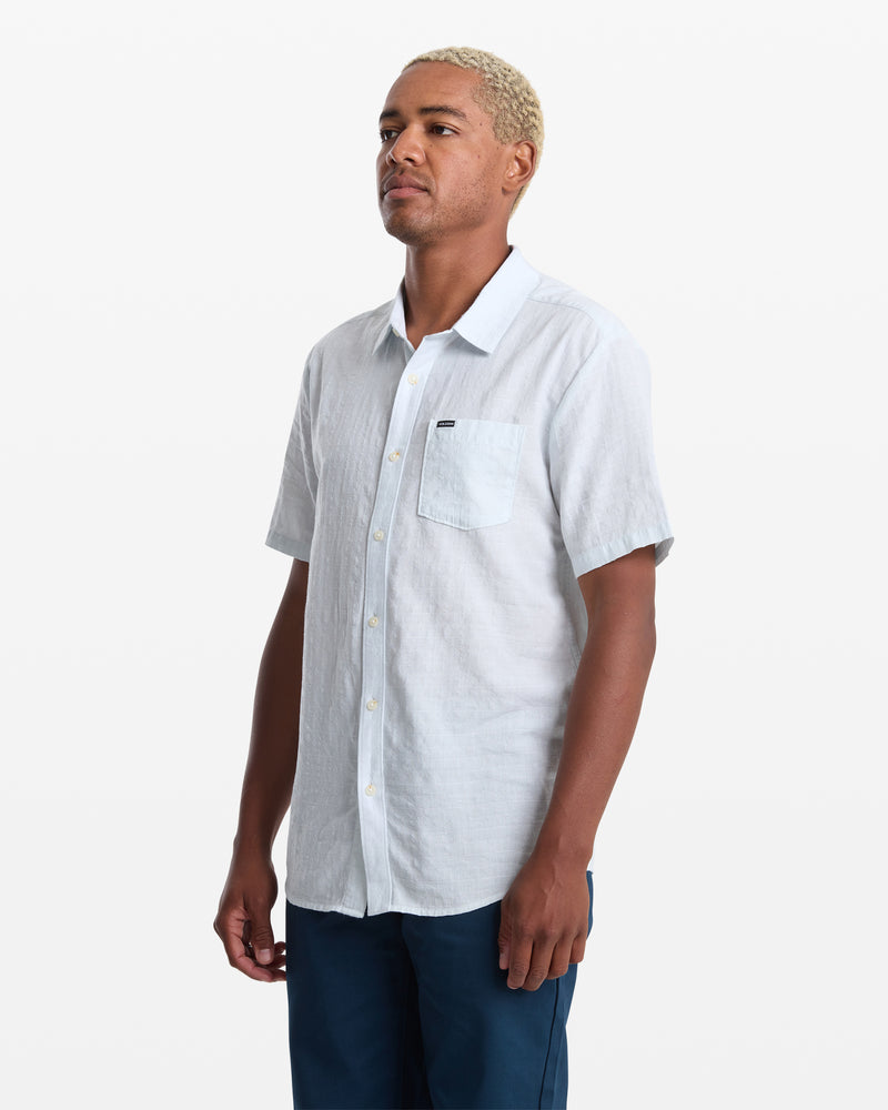 A person with short blond hair wears the Volcom Men's Bankstone II Short Sleeve shirt in light blue and dark blue pants, standing against a plain white background.