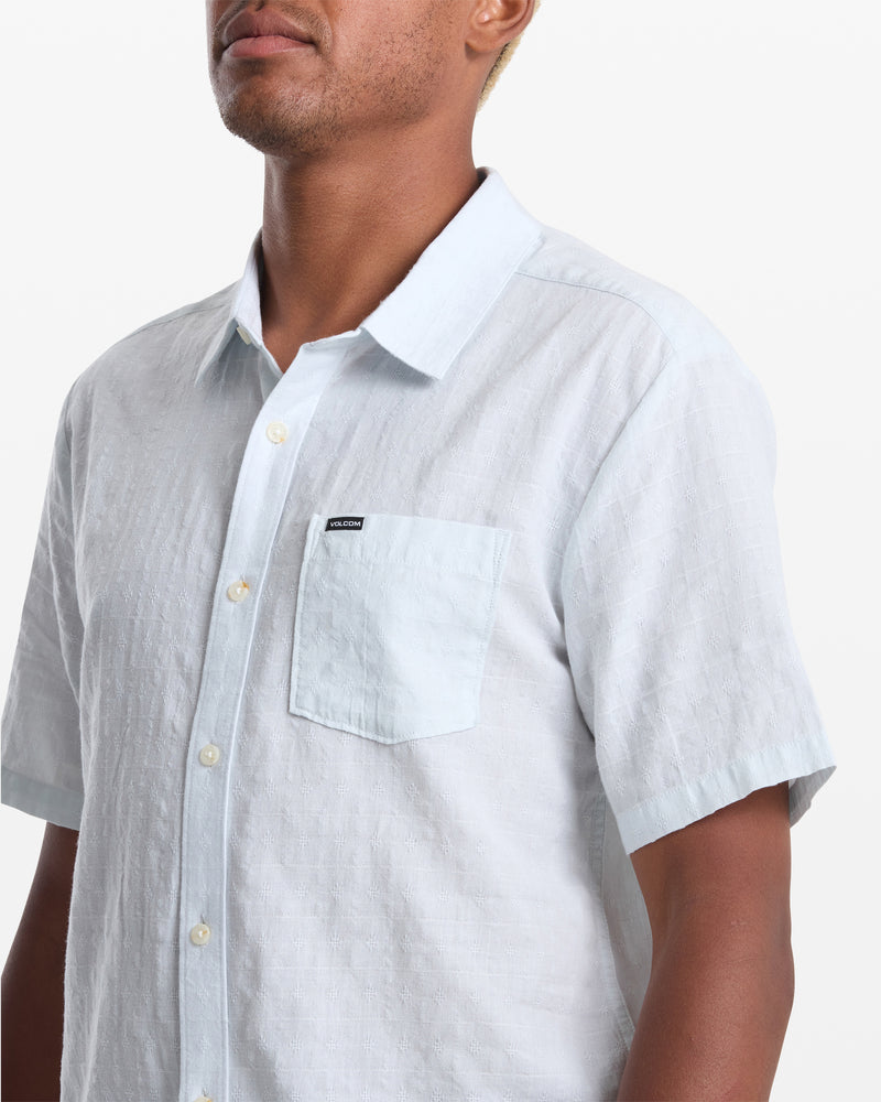 A man wears the Volcom Men's Bankstone II Short Sleeve—a light blue, short-sleeve button-up with a front pocket and black label—shown from shoulders to mid-torso against a plain white background.