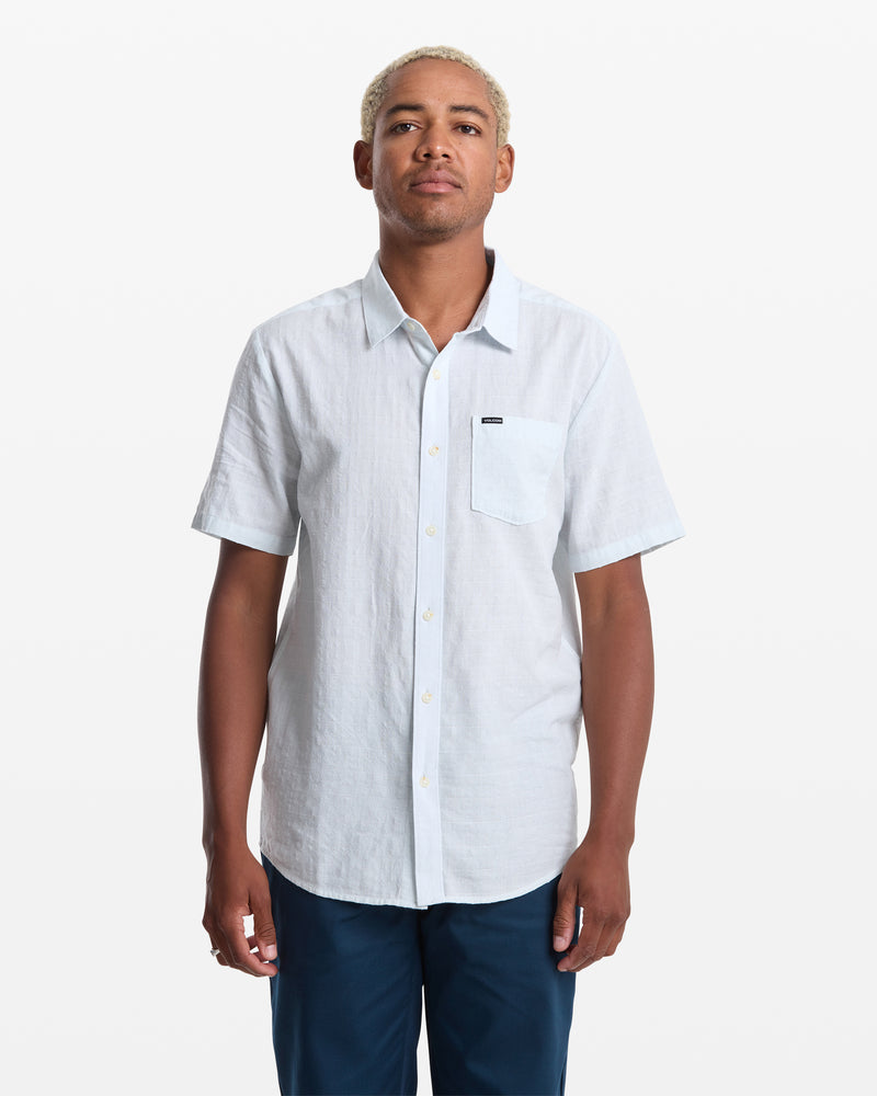A man with short, light blond hair models the Volcom Men's Bankstone II Short Sleeve button-up shirt in light blue, featuring a left chest pocket, against a plain white background.