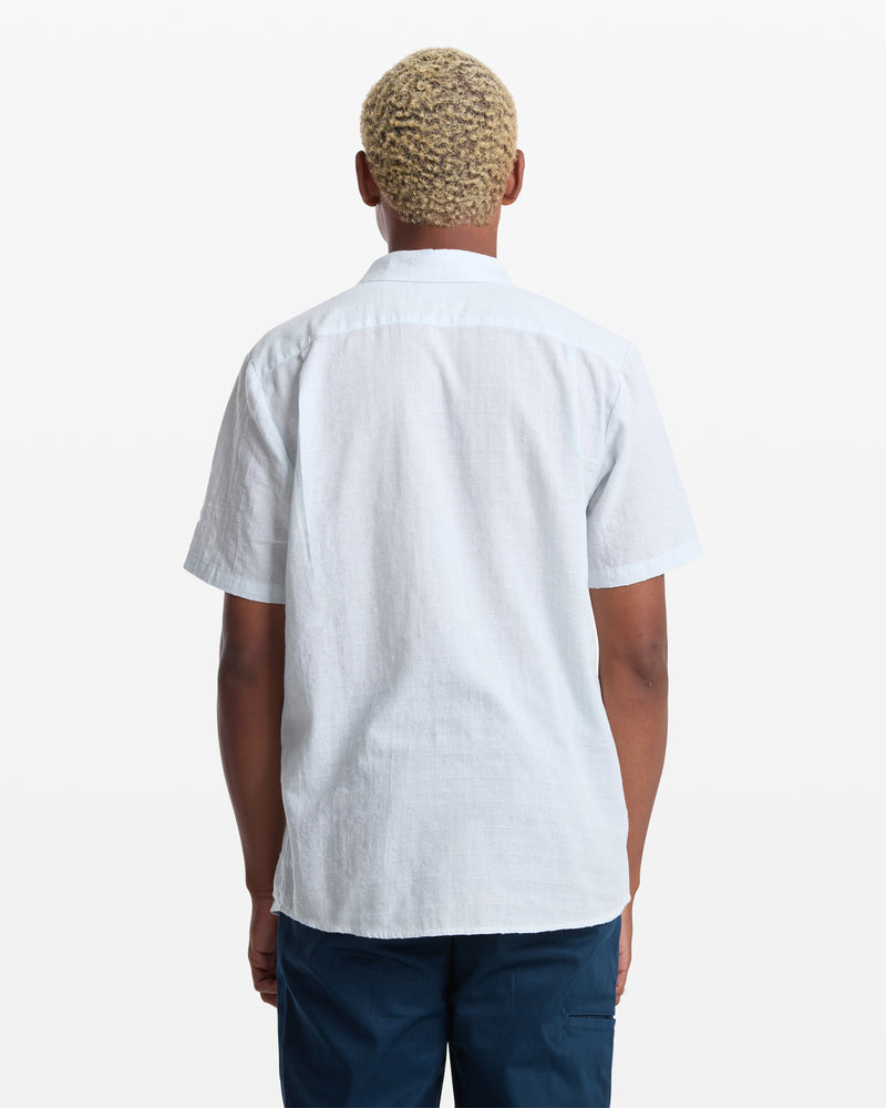 A person with short, bleached hair is shown from the back wearing the Volcom Men's Bankstone II Short Sleeve shirt in light blue and dark blue pants against a plain white background.