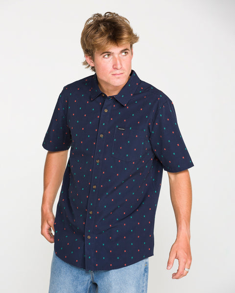 A person with light brown hair wears the Volcom Men's Bankstone Short Sleeve Shirt in a classic fit, featuring a subtle dobby weave pattern in navy, paired with light blue jeans and standing against a plain white background.