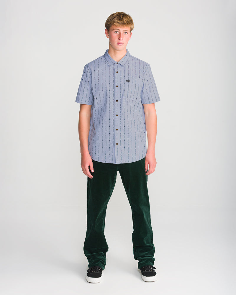 A person wearing a pale blue, short-sleeved, pinstriped Volcom Men’s Bankstone Short Sleeve shirt, dark green pants, and black sneakers.