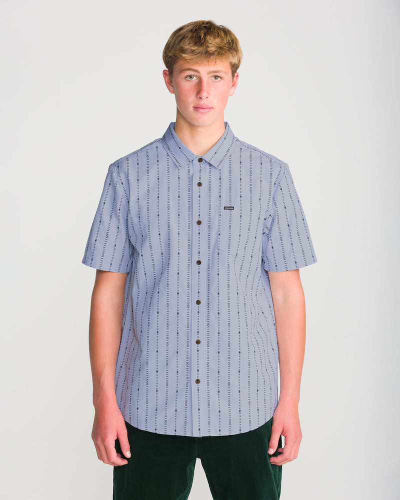 A young man wears a Volcom Men’s Bankstone Short Sleeve, a pale blue button-down shirt featuring a vertical dashed-line pattern and a single chest pocket.