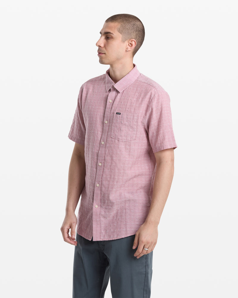 A man stands against a plain white background wearing the Volcom Men's Bankstone II Short Sleeve shirt in light pink, featuring a chest pocket, and dark gray pants. He faces slightly to the side with a neutral expression.
