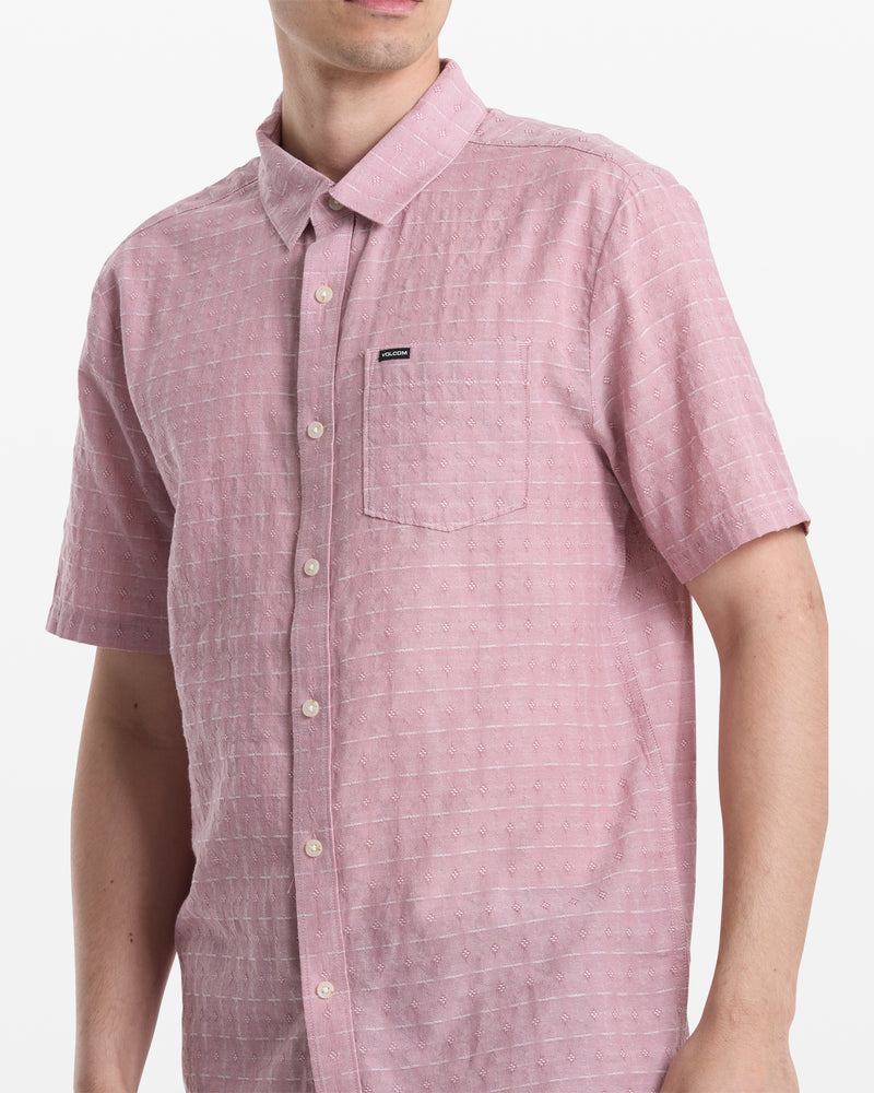 A man wears the Volcom Men's Bankstone II Short Sleeve—a light pink, checkered button-up with a left chest pocket—standing against a plain white background.