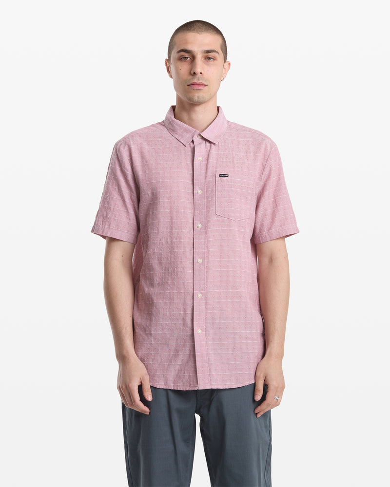 A man with short hair models the Volcom Men's Bankstone II Short Sleeve pink button-up shirt with a chest pocket and gray pants, standing against a plain white background.