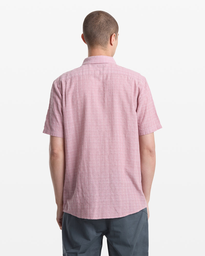 A man with short hair, seen from behind, wears the Volcom Men's Bankstone II Short Sleeve in light pink and dark gray pants, standing against a plain white background.