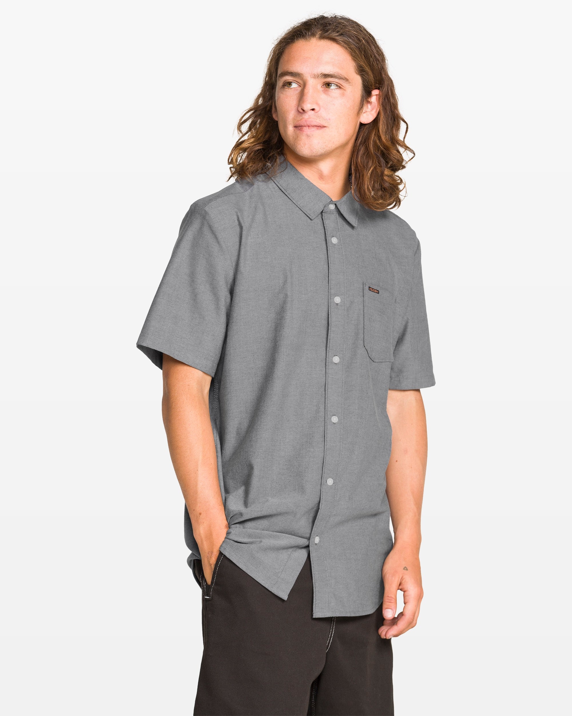 A young man with long brown hair, dressed in Volcom's Men's Everett Oxford II shirt and black pants, stands against a plain white background, gazing off to the side with a relaxed expression.