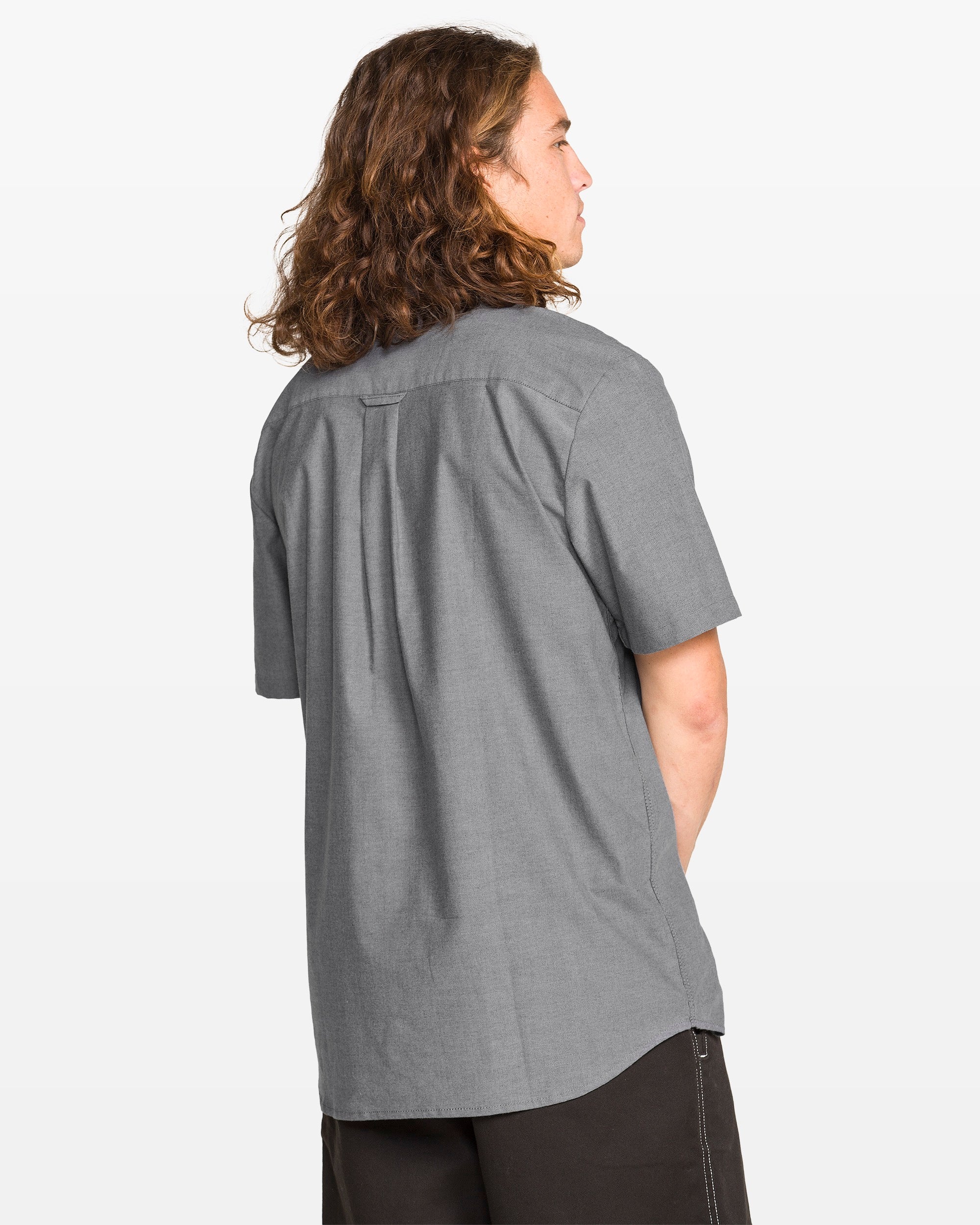 A person with long brown hair is shown from the back wearing a light gray Men's Everett Oxford II short sleeve shirt by Volcom and dark shorts, set against a plain white background.