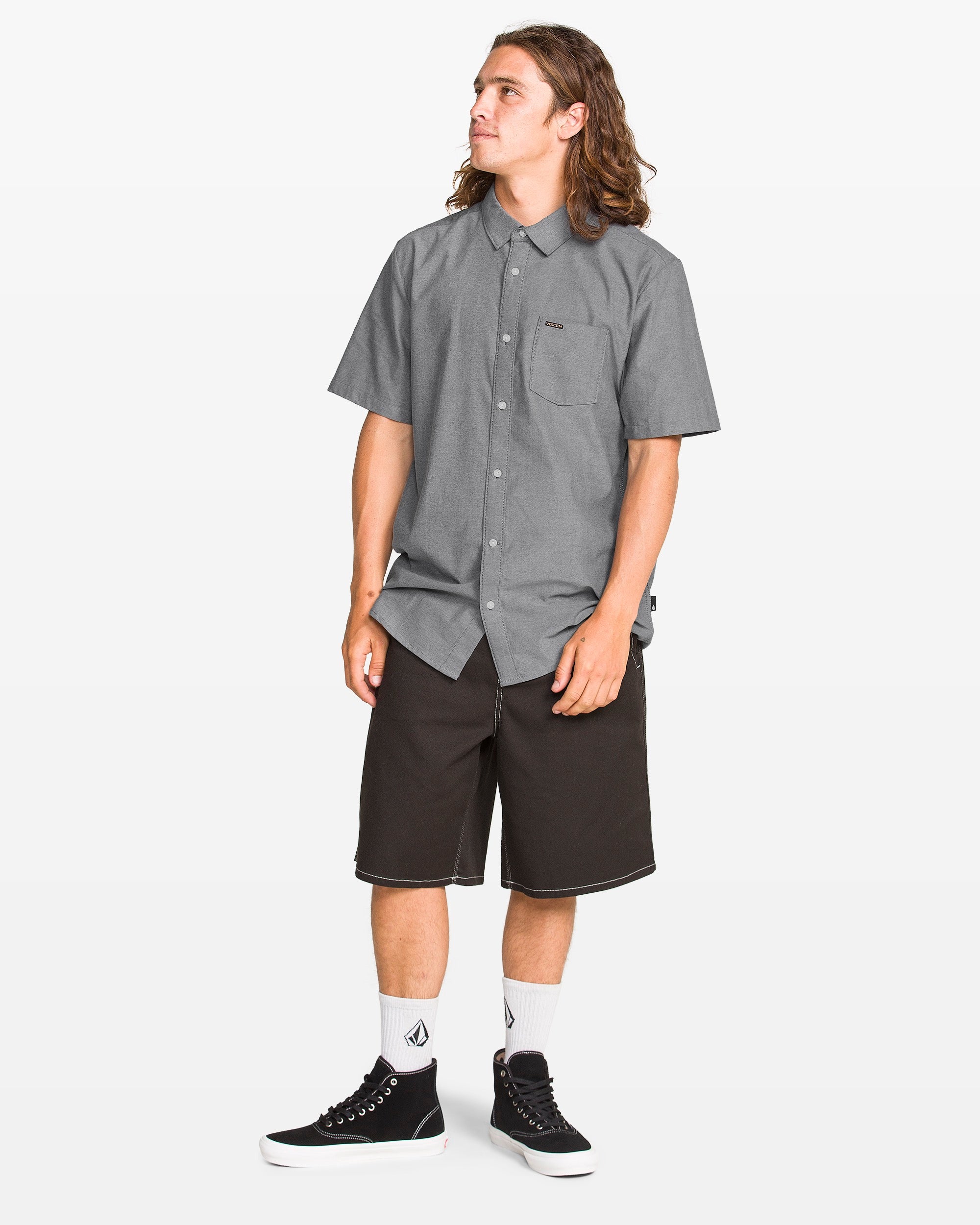 A person with long hair wears the Volcom Men's Everett Oxford II shirt in gray, black knee-length shorts, white logo socks, and black sneakers while standing on a plain white background and looking to the side.
