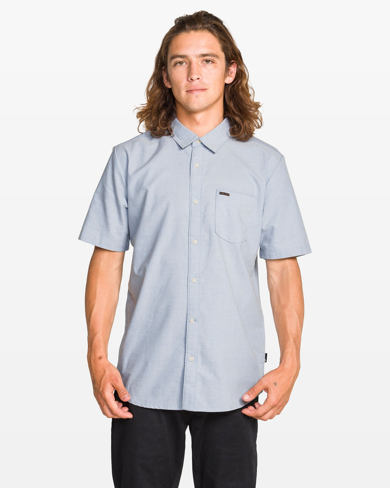 A young man with long brown hair wears a Volcom Men's Everett Oxford II Shirt in light blue and black pants, standing facing forward against a white background to showcase its classic fit.