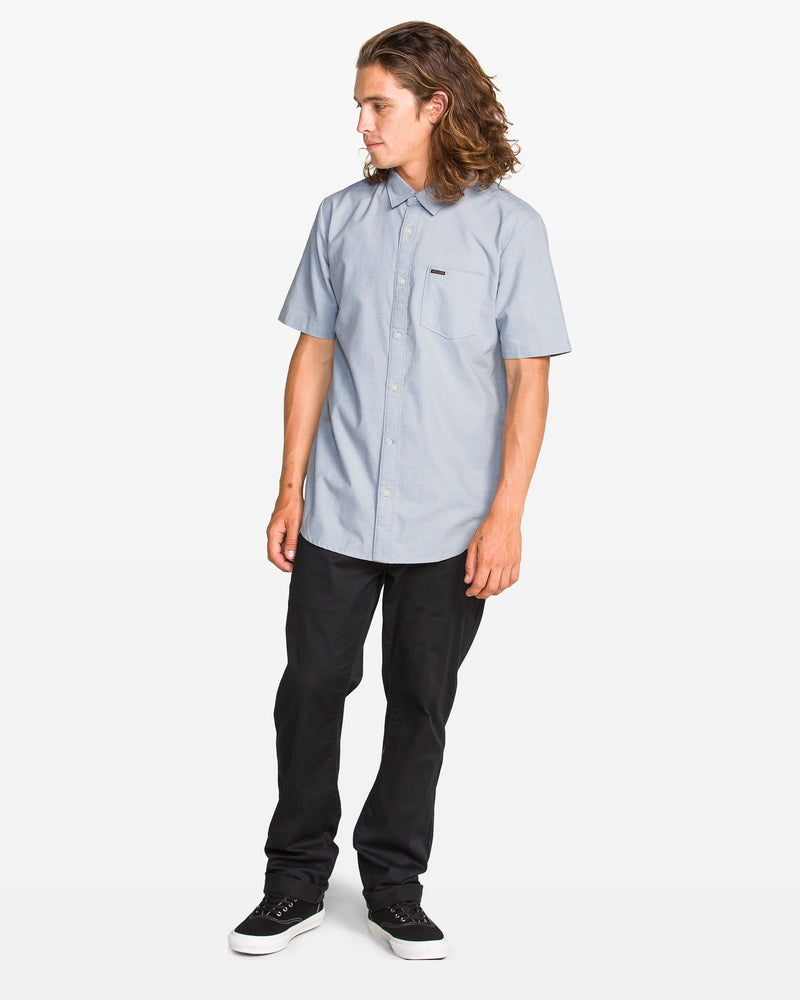 A person with long brown hair wears a Volcom Men's Everett Oxford II Shirt in light blue, classic fit black pants, and black sneakers while standing and looking to the side against a plain white background.