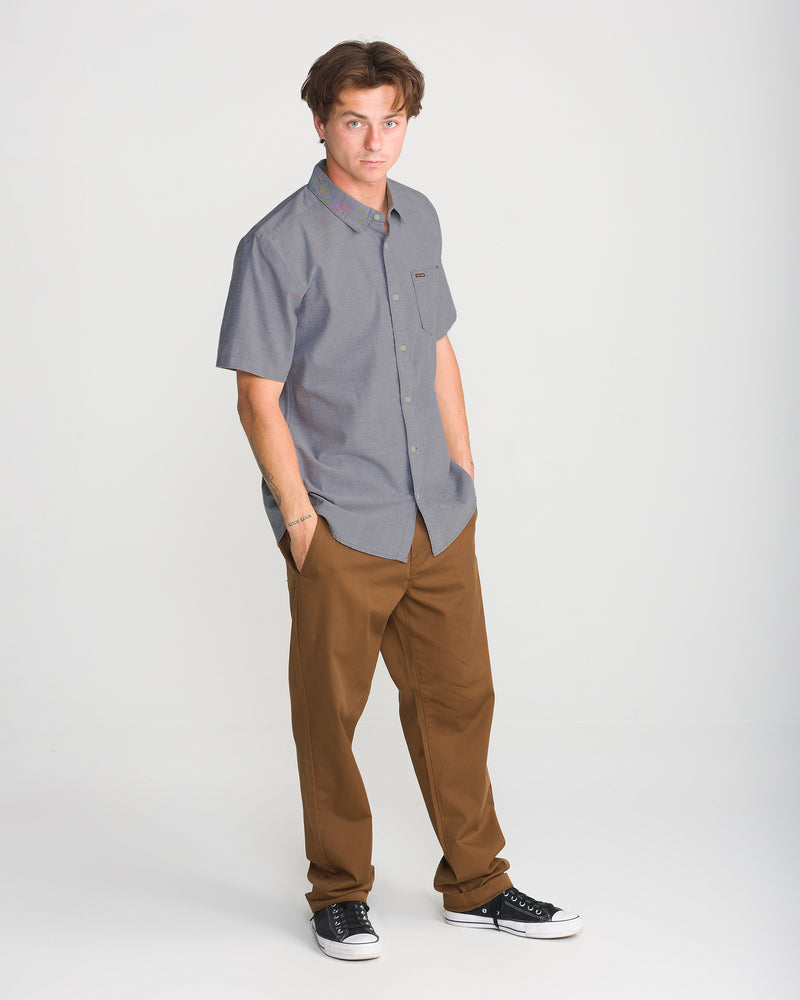 A man wears a gray Volcom Men’s Everett Oxford II button-up shirt with brown trousers and black low-top sneakers.