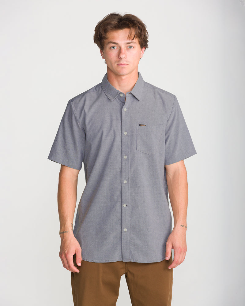 A man wearing a gray Volcom Men’s Everett Oxford II stretch button-up shirt with a chest pocket and brown pants.
