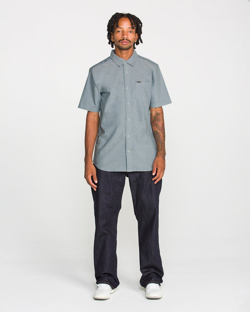 A man with short dreadlocks wears the Volcom Men's Everett Oxford II Short Sleeve Shirt in grey, styled with dark jeans and white sneakers, standing against a plain white background. This shirt’s stretch-woven fabric provides comfortable wear while maintaining a relaxed yet polished look.