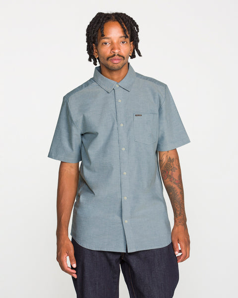 A man with short dreadlocks and a tattooed arm wears the Volcom Men's Everett Oxford II Short Sleeve Shirt—a classic-fit, button-up style with a convenient front pocket, crafted from stretch-woven fabric for extra comfort. He stands against a plain white background.