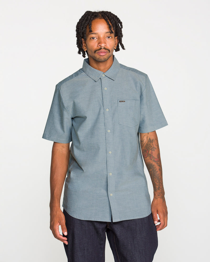A man with short dreadlocks and a tattooed arm wears the Volcom Men's Everett Oxford II Short Sleeve Shirt—a classic-fit, button-up style with a convenient front pocket, crafted from stretch-woven fabric for extra comfort. He stands against a plain white background.