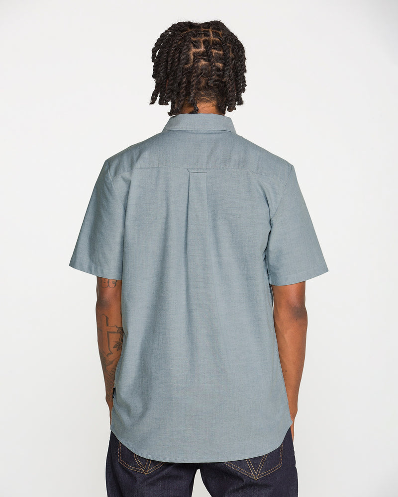 A person with short dreadlocks, wearing a light blue Volcom Men's Everett Oxford II Short Sleeve Shirt and dark jeans, is standing with their back to the camera against a plain white background.