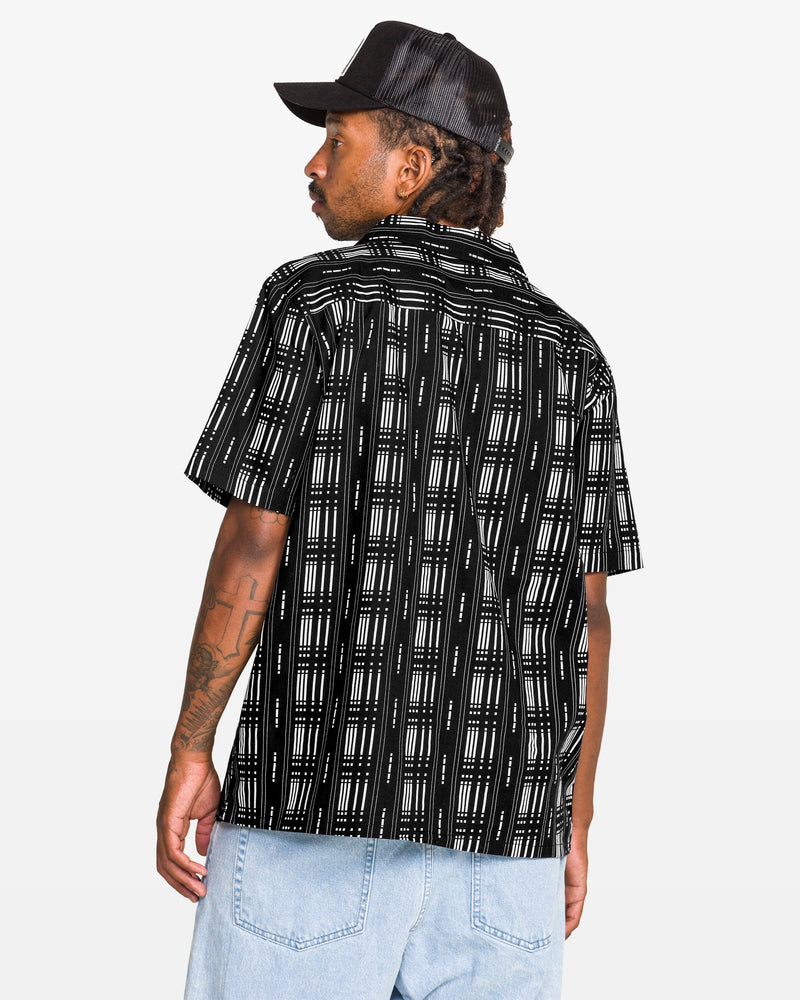 Man wearing the Volcom Men's Macros Short Sleeve Tee, featuring a black allover print and boxy fit, paired with light blue jeans and a black cap, facing away over his shoulder against a plain white background.