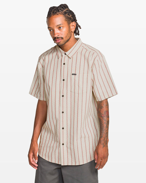 A man with short dreadlocks and tattoos on his left arm wears the Volcom Men's Sato Stripe Short Sleeve Tee, featuring yarn-dyed vertical red stripes, along with dark pants, standing against a plain white background.