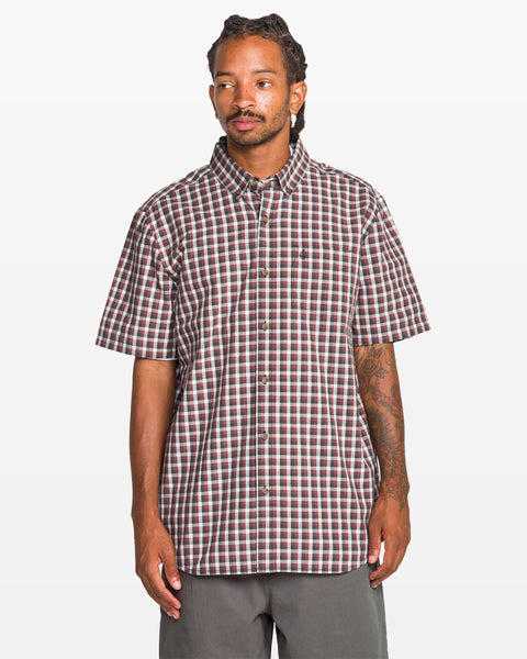 A man with long braided hair and a tattooed right arm wears a Volcom Men's Louie Check Short Sleeve Tee in light blue acid check with gray pants, standing against a plain white background.