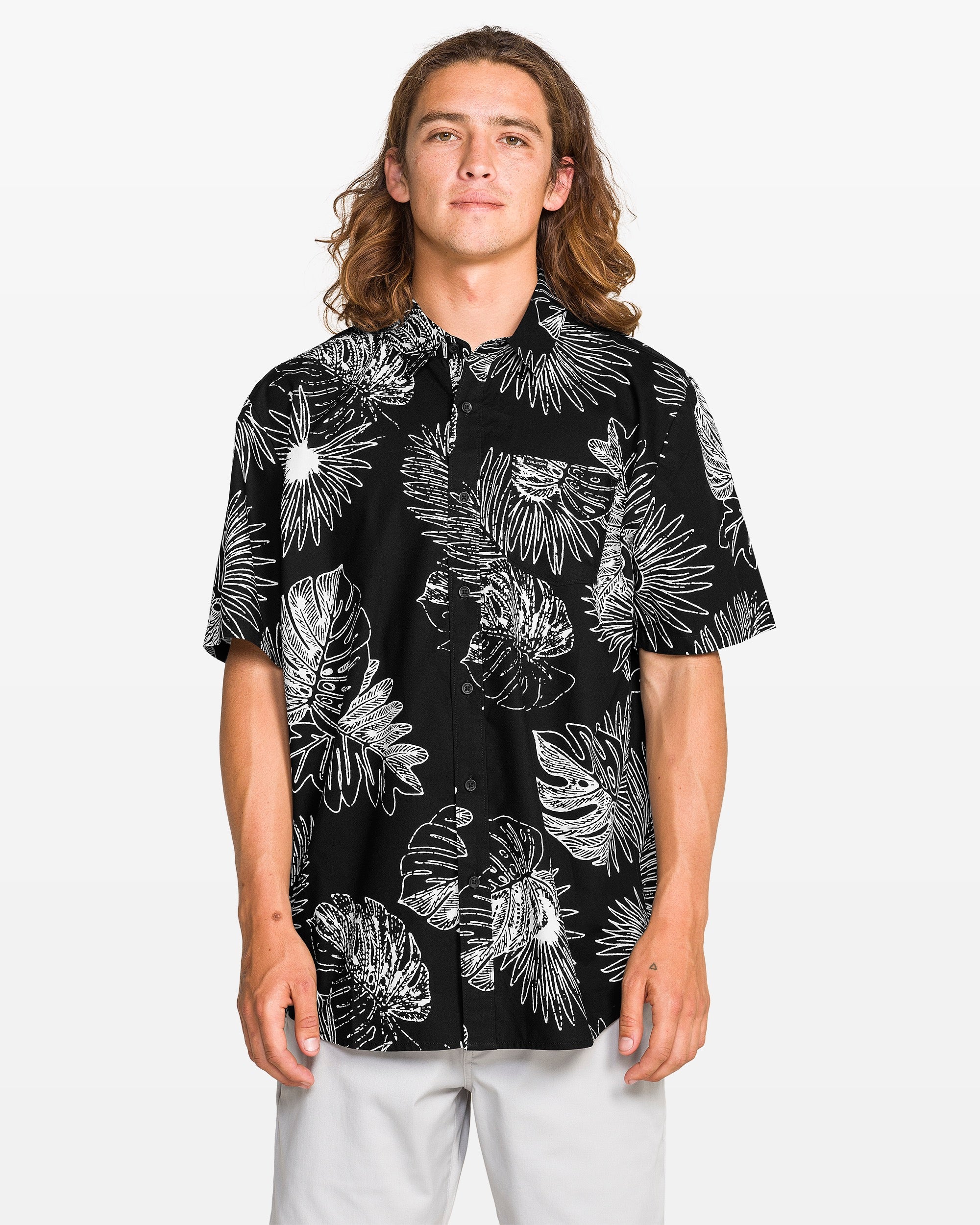 A man with long, wavy brown hair wears the Volcom Men's Monsterious Short Sleeve Tee, featuring an all-over print of white tropical leaves, paired with light pants and standing against a plain light background.