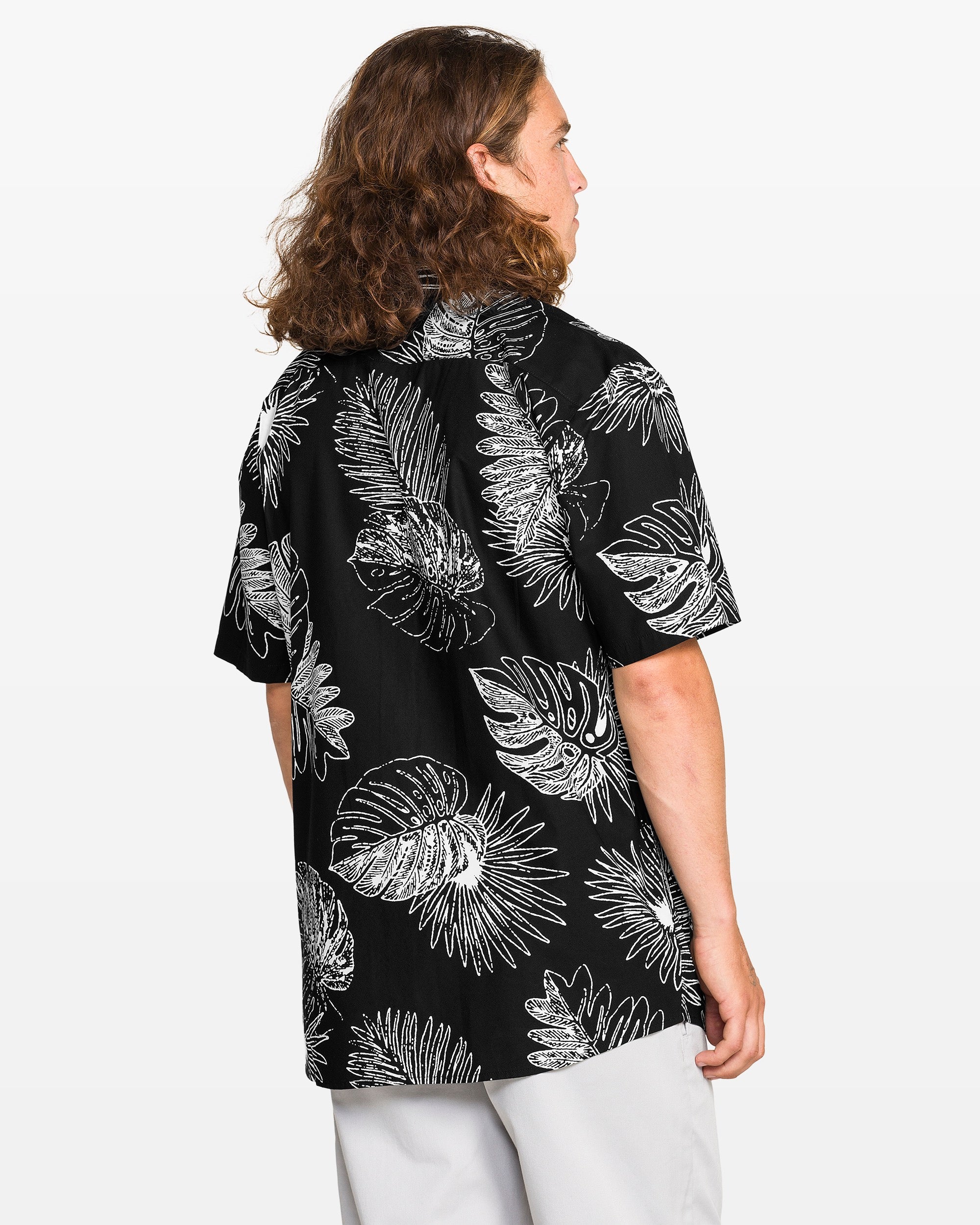 A person with long brown hair is seen from the back wearing Volcom’s Men's Monsterious Short Sleeve Tee, a black classic-fit shirt with white tropical leaf print, paired with light-colored pants against a plain background.