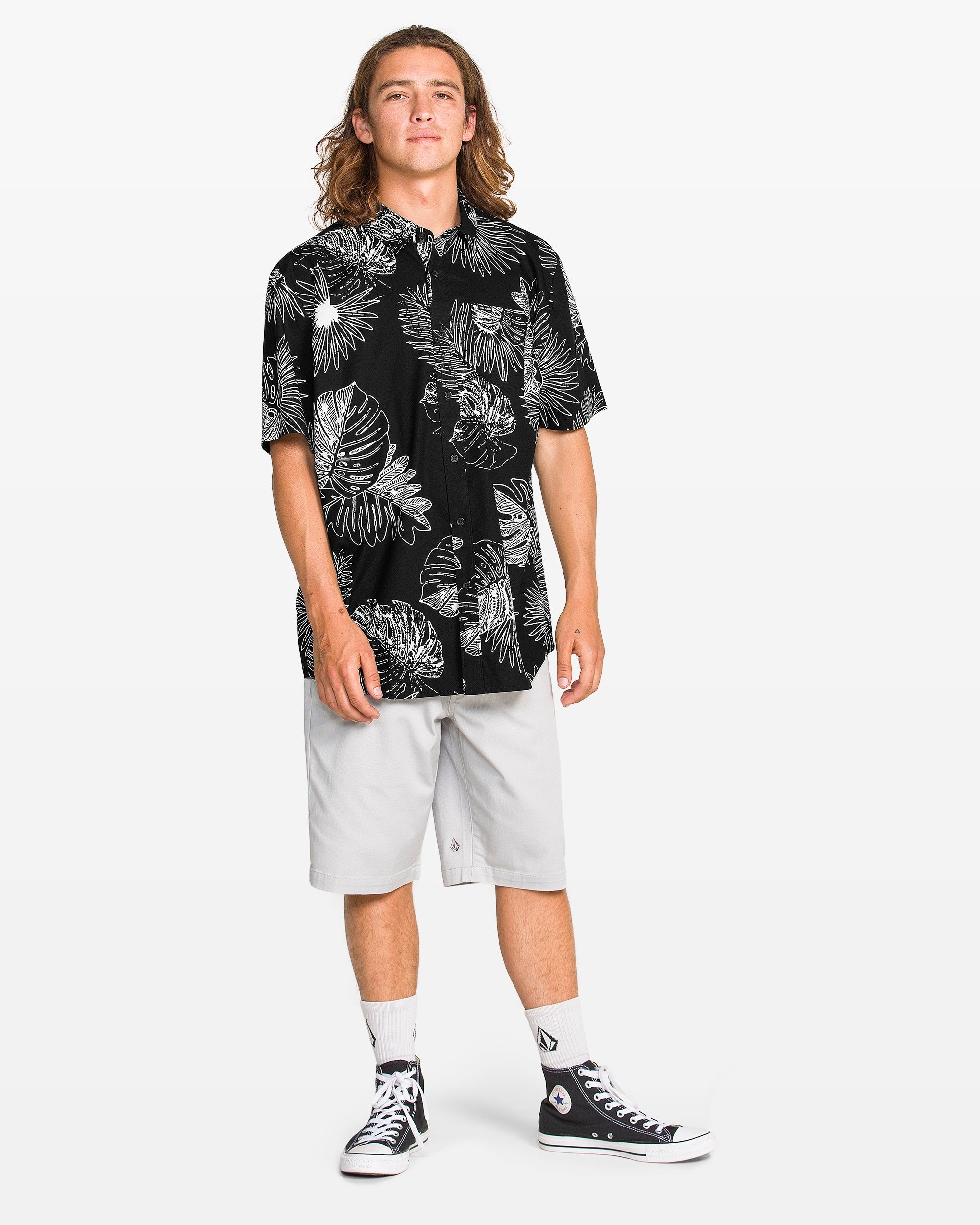 A young person with long hair wears a Volcom Men's Monsterious Short Sleeve Tee in a classic fit with all-over white tropical leaf print, light gray knee-length shorts, white crew socks, and black high-top sneakers against a plain white background.