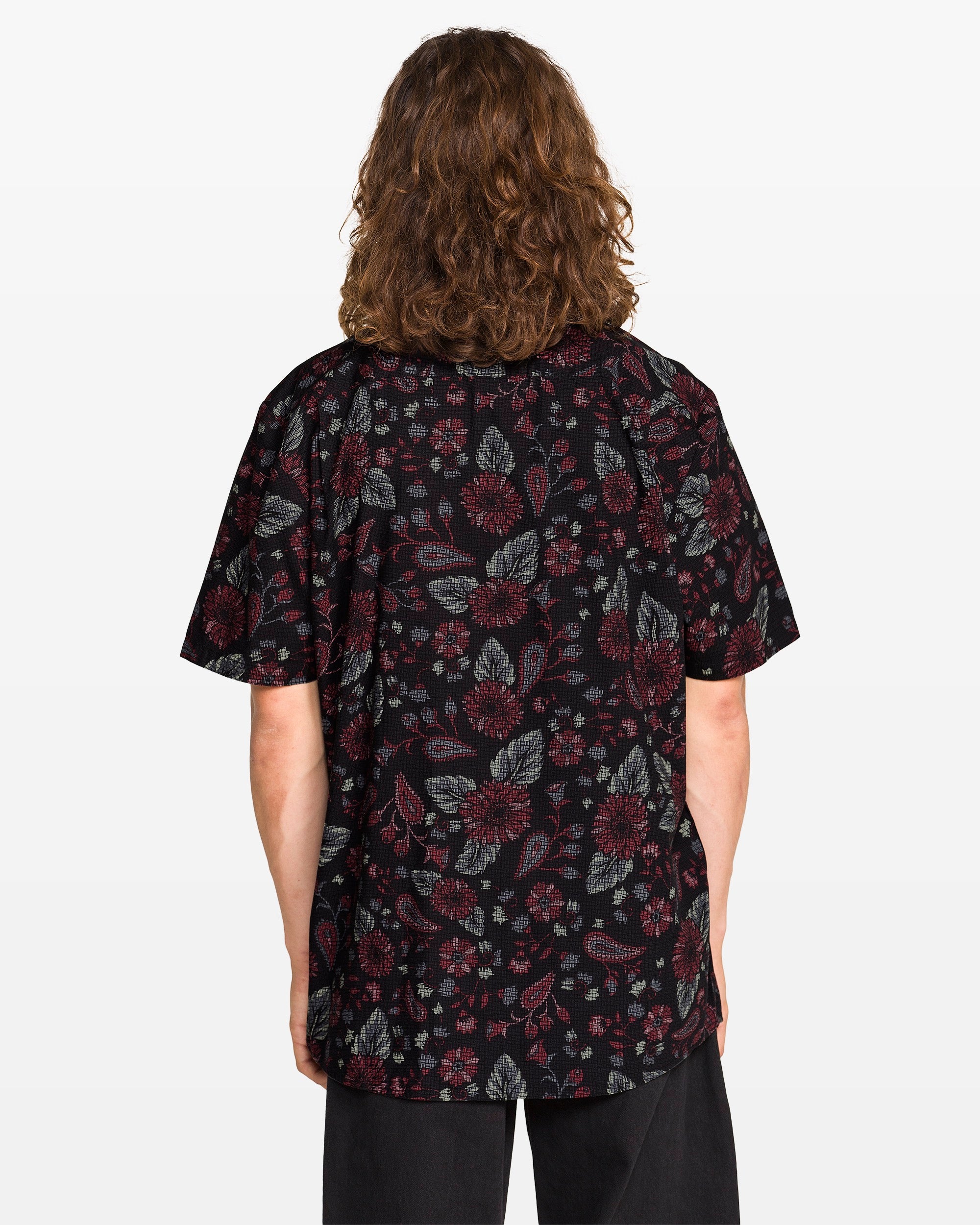 A man with long brown hair is seen from behind wearing the Volcom Men's Scaler Stone Woven Short Sleeve Shirt in black, featuring a red and gray floral pattern, against a plain white background.