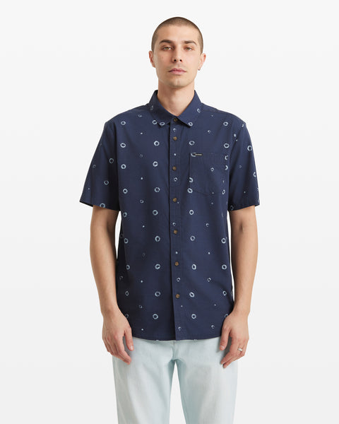 A person stands facing forward against a plain white background, wearing the Volcom Men's Scaler Stone Woven Short Sleeve Shirt—an allover circular dot print shirt in navy blue—with light-colored pants.