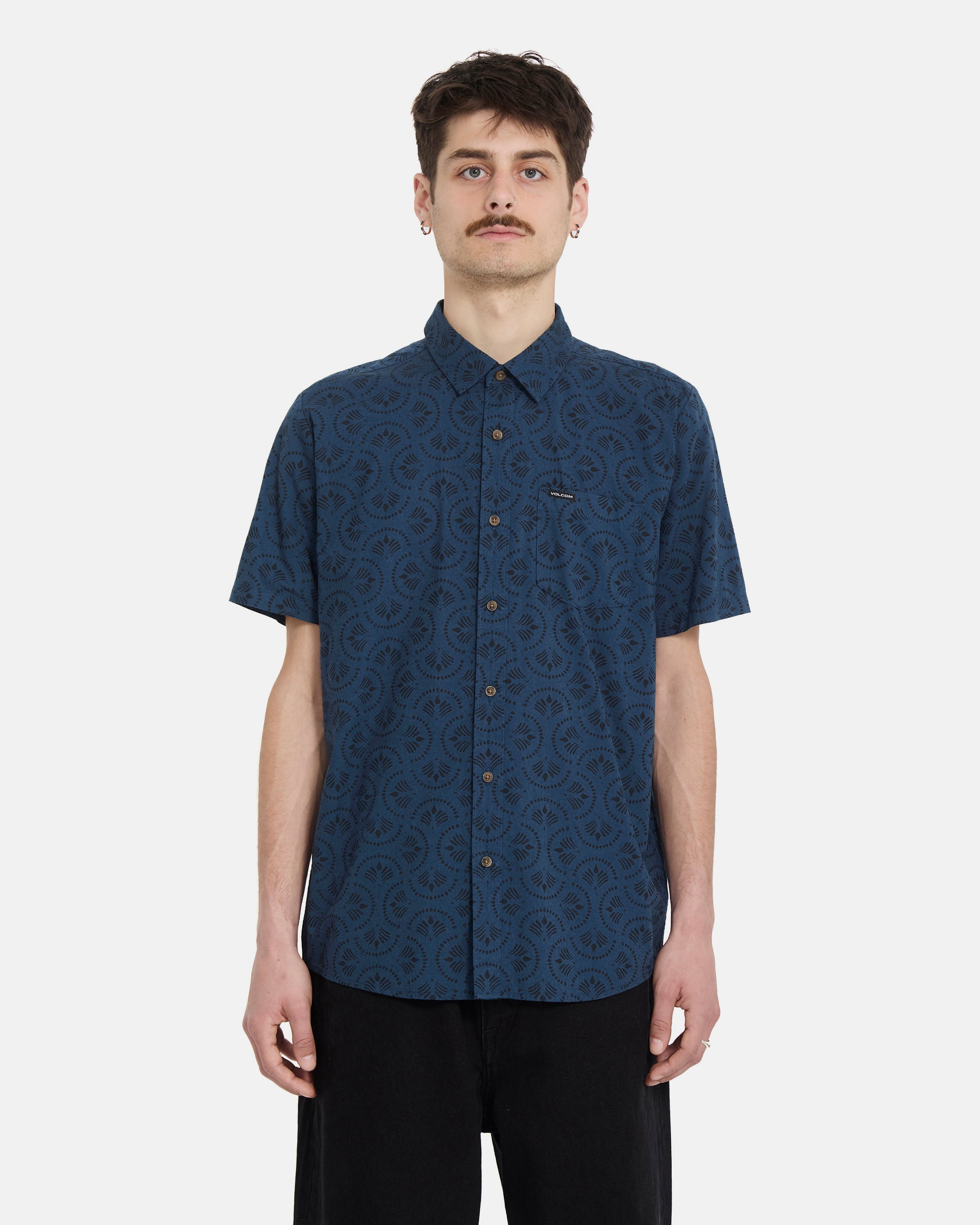 A young man with short dark hair and a mustache wears the Volcom Men's Scaler Stone Woven Short Sleeve Shirt in midnight blue with black pants, standing facing forward against a plain white background.