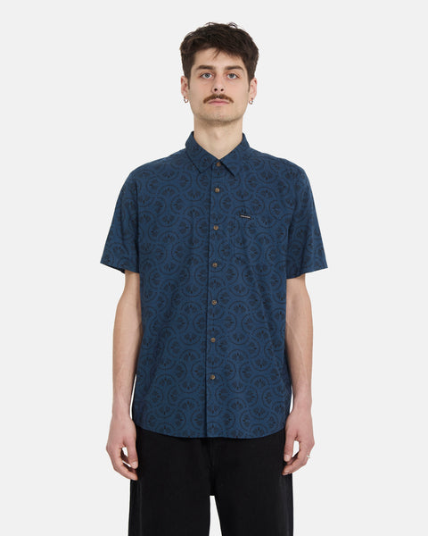 A young man with short dark hair and a mustache wears the Volcom Men's Scaler Stone Woven Short Sleeve Shirt in midnight blue with black pants, standing facing forward against a plain white background.