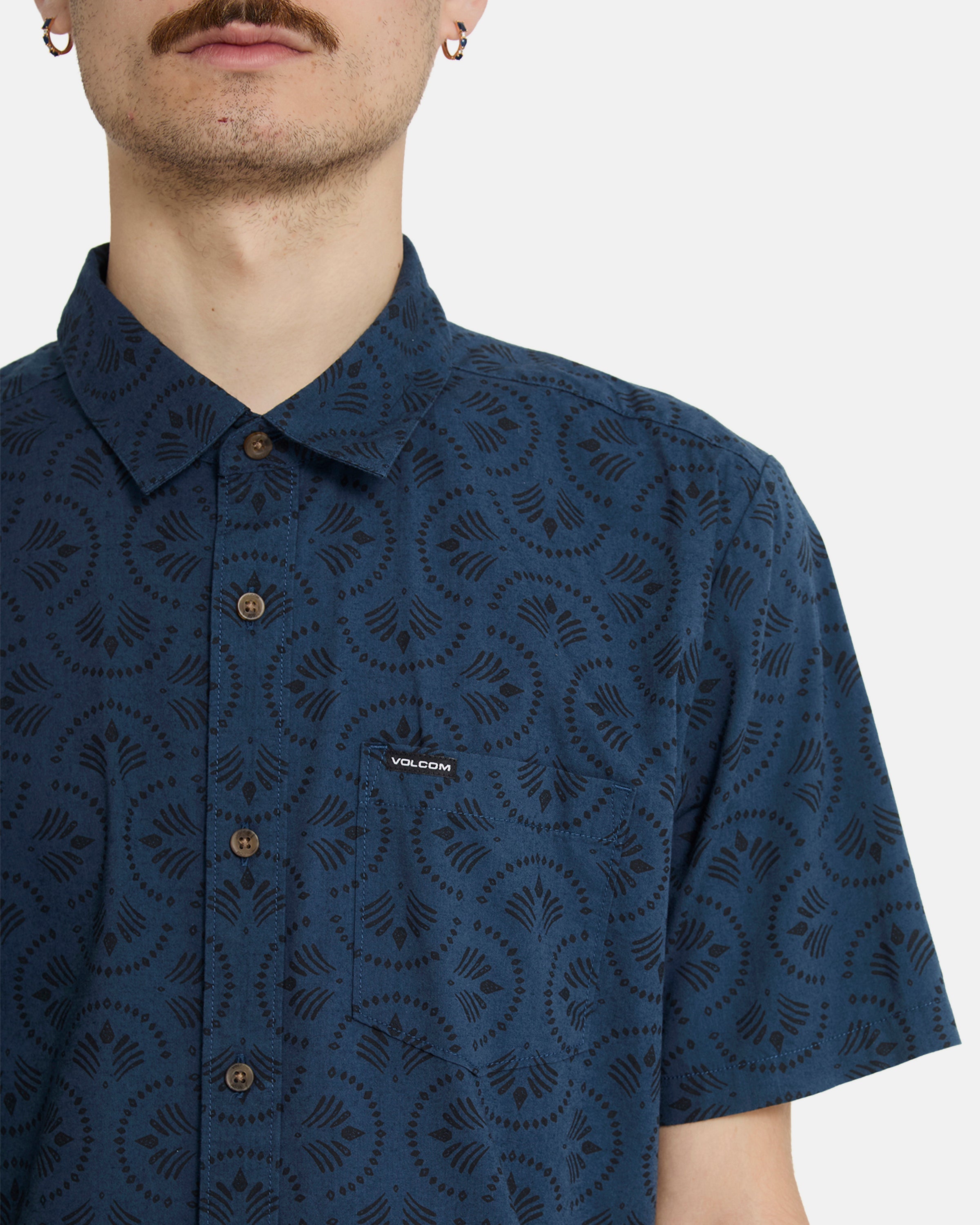 A person wears the Volcom Men's Scaler Stone Woven Short Sleeve Shirt in midnight blue with a black geometric pattern and Volcom logo on the chest pocket; the image is cropped from nose to just below the chest.