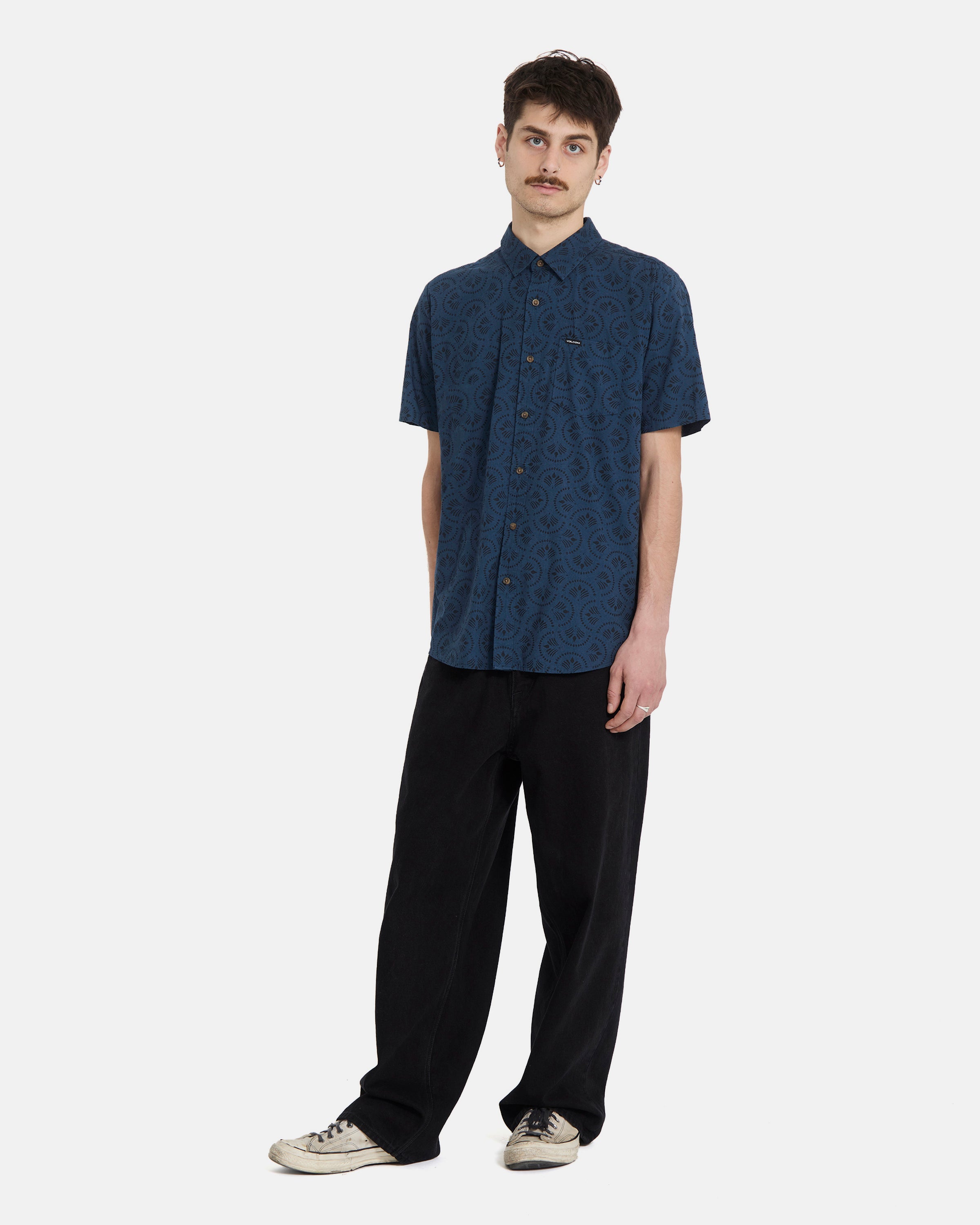 A man with short brown hair and a mustache wears the Volcom Men's Scaler Stone Woven Short Sleeve Shirt in midnight blue, paired with loose black pants and white sneakers, standing against a plain white background.
