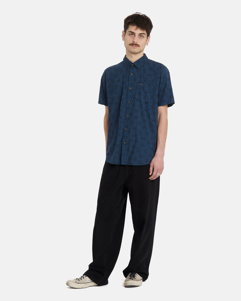 A man with short brown hair and a mustache wears the Volcom Men's Scaler Stone Woven Short Sleeve Shirt in midnight blue, paired with loose black pants and white sneakers, standing against a plain white background.