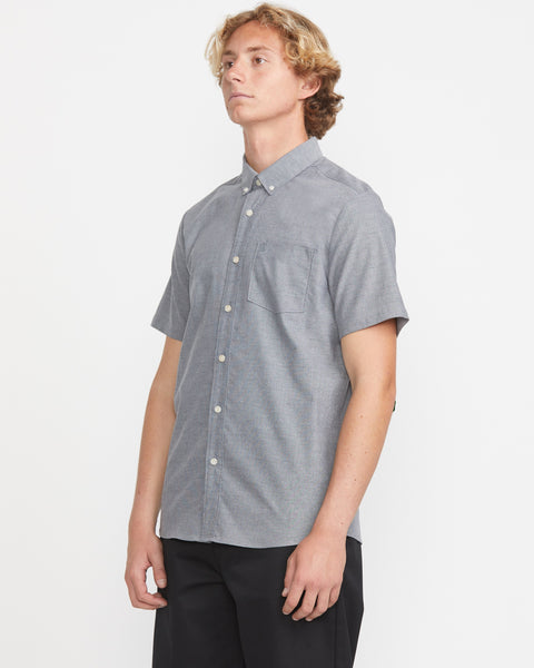 A person with short blond hair wears the Volcom Men's Everett Oxford Short Sleeve Tee, which features branded details and a chest pocket, paired with black pants, standing against a plain white background.